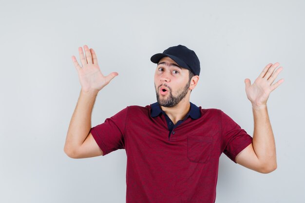 Jeune homme en t-shirt rouge, casquette noire montrant un geste impuissant et regardant choqué, vue de face.