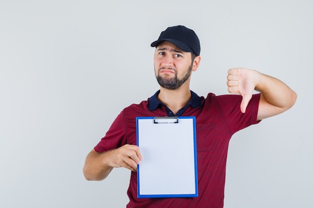 Jeune homme en t-shirt rouge, casquette noire montrant le pouce vers le bas tout en tenant le cahier et à la triste, vue de face.