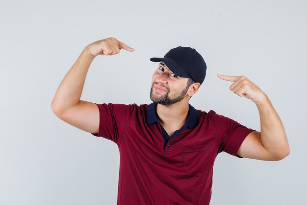 Jeune homme en t-shirt rouge, casquette noire pointant sur sa casquette et à la jolie vue de face.