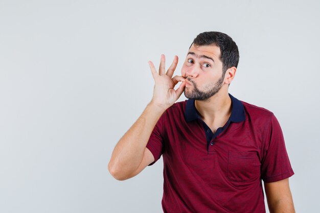 Jeune homme en t-shirt rouge montrant le geste de silence et à la vue de face, focalisée.