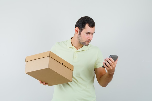 Jeune homme en t-shirt tenant une boîte en carton, regardant la calculatrice, vue de face.