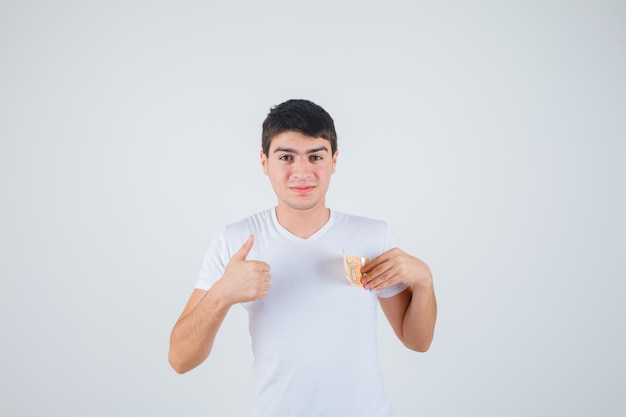 Jeune homme en t-shirt tenant eurobanknote, montrant le pouce vers le haut et l'air heureux, vue de face.