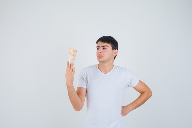 Jeune homme en t-shirt tenant eurobanknote, regardant eurobanknote et regardant focalisé, vue de face.