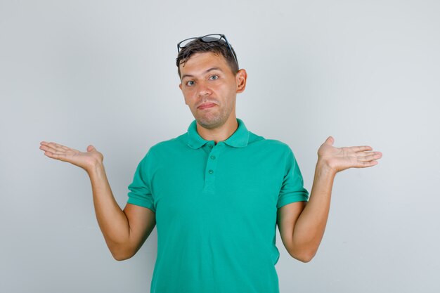 Jeune homme en t-shirt vert, lunettes montrant un geste impuissant avec les mains, vue de face.