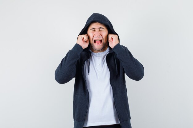 Jeune homme en t-shirt, veste bouchant les oreilles avec les doigts et à l'ennui, vue de face.