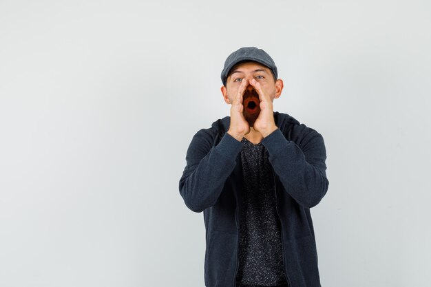 Jeune homme en t-shirt, veste, casquette disant secret avec les mains près de la bouche, vue de face.