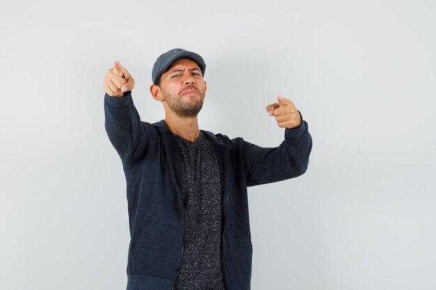 Jeune homme en t-shirt, veste, casquette pointant vers la caméra, vue de face.