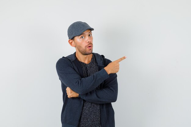 Jeune homme en t-shirt, veste, casquette pointant vers l'extérieur et regardant curieux, vue de face.