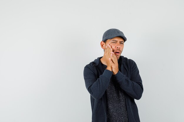 Jeune homme en t-shirt, veste, casquette souffrant de maux de dents douloureux et à la vue inconfortable, de face.