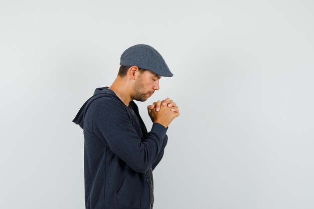 Jeune homme en t-shirt, veste, casquette tenant les mains jointes en geste de prière et à la recherche de calme.