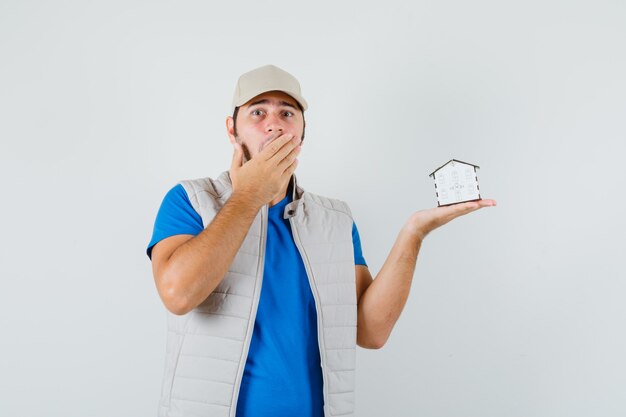 Jeune homme en t-shirt, veste, casquette tenant le modèle de la maison et à la surprise, vue de face.