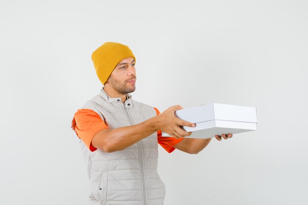 Jeune homme en t-shirt, veste, chapeau livrant une boîte en carton et à la douce vue de face.