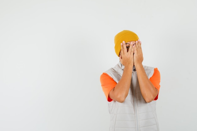 Jeune homme en t-shirt, veste, chapeau regardant à travers les doigts et à la peur