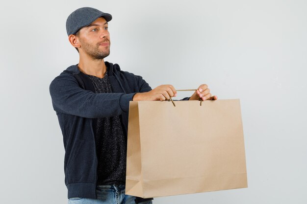 Jeune homme en t-shirt, veste, jeans, casquette livrant un sac en papier, vue de face.