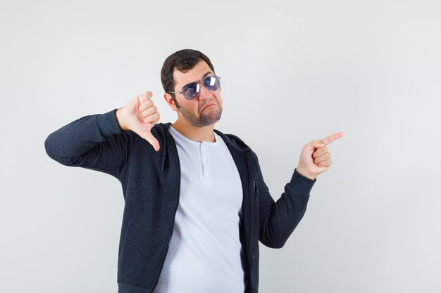 Jeune homme en t-shirt, veste pointant sur le côté, montrant le pouce vers le bas et regardant hésitant, vue de face.