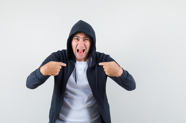 Jeune homme en t-shirt, veste pointant sur lui-même tout en criant et à la vue agressive, de face.