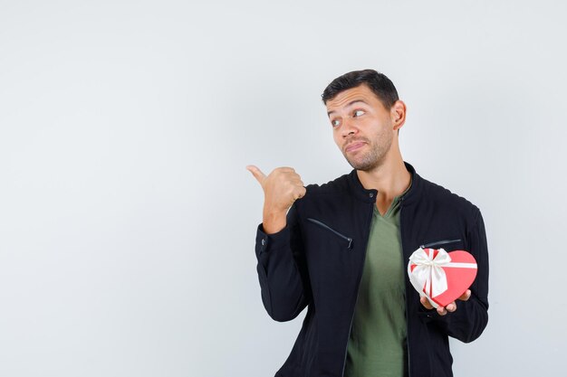 Jeune homme en t-shirt, veste pointant le pouce vers l'extérieur, tenant une boîte-cadeau et l'air positif, vue de face.