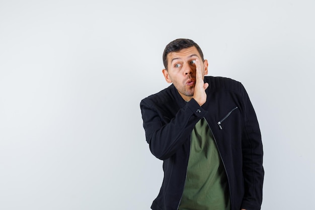 Jeune homme en t-shirt, veste racontant le secret derrière la main et l'air curieux, vue de face.