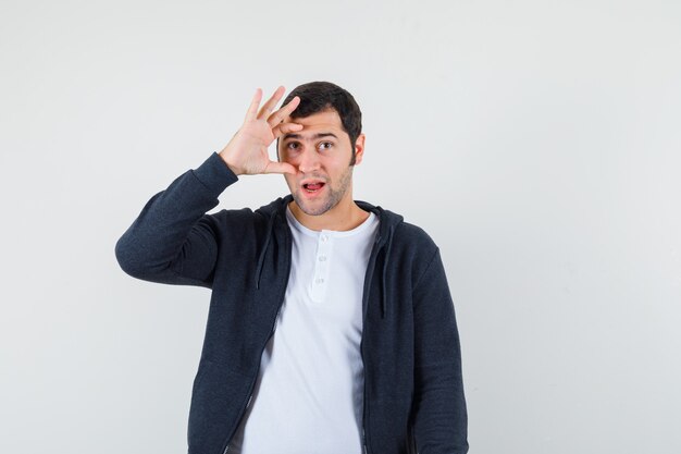 Jeune homme en t-shirt, veste regardant à travers les doigts et à la vue de face, confiant.