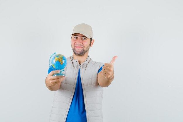 Jeune homme en t-shirt, veste tenant le globe de l'école, montrant le pouce vers le haut et à la joyeuse vue de face.