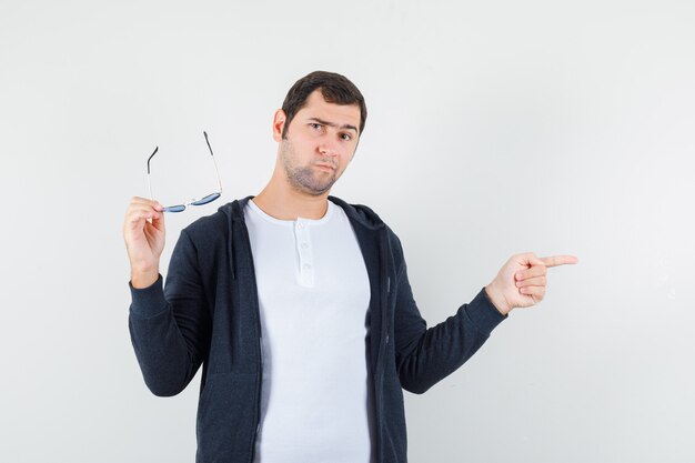 Jeune homme en t-shirt, veste tenant des lunettes, pointant de côté et regardant hésitant, vue de face.