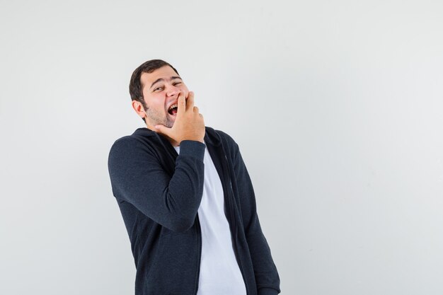 Jeune homme en t-shirt, veste tenant la main sur la bouche en riant, vue de face.