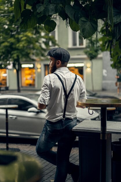 Jeune homme tatoué barbu dans un café dans la rue avec un verre de vin. Un mec romantique avec une casquette blanche et des bretelles dans la ville. Peaky Blinders. vieux rétro à la mode.