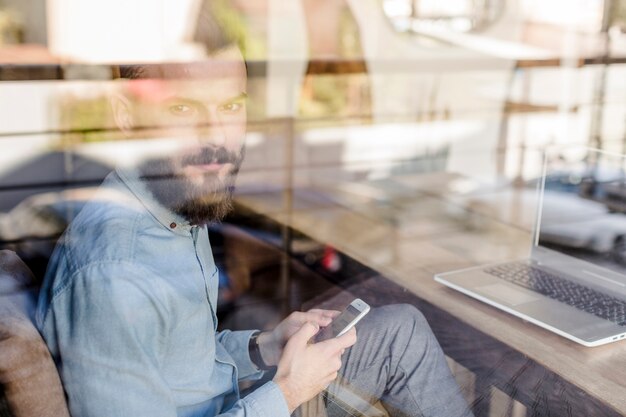 Jeune homme avec un téléphone portable vu à travers un verre transparent dans un café