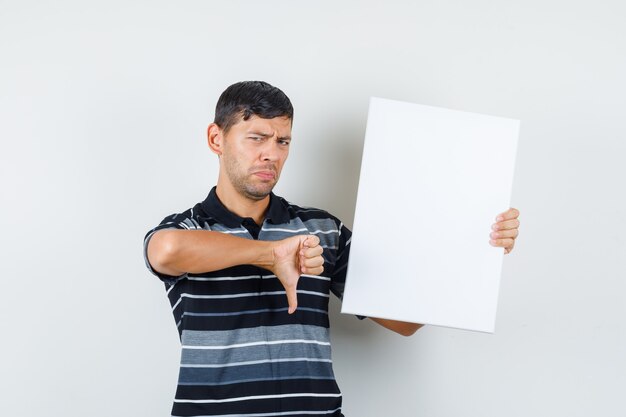 Jeune homme tenant une affiche vierge avec le pouce vers le bas en t-shirt et à la vue de face, modérée.