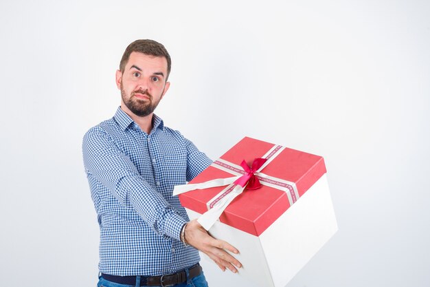 Jeune homme tenant une boîte-cadeau en chemise et à la perplexité. vue de face.