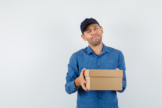 Jeune homme tenant une boîte en carton en chemise bleue, une casquette et à la recherche désespérée, vue de face.