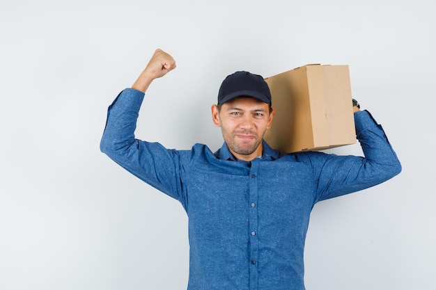 Jeune Homme Tenant Une Boîte En Carton Avec Le Geste Du Gagnant En Chemise Bleue, Casquette Et L'air Chanceux, Vue De Face.