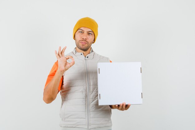 Jeune homme tenant une boîte en carton, montrant le geste ok en t-shirt, veste, chapeau et à la jolly. vue de face.