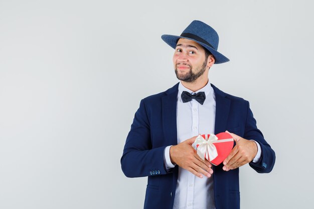 Jeune homme tenant la boîte présente en costume, chapeau et à la joyeuse vue de face.