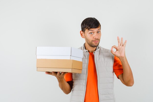 Jeune homme tenant des boîtes en carton, montrant le geste ok en t-shirt, vue de face de la veste.