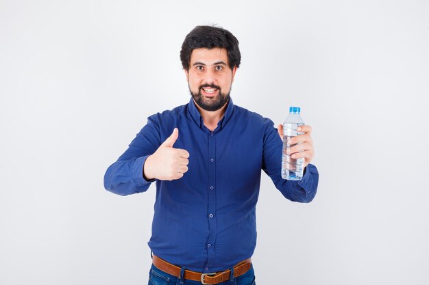 Jeune homme tenant une bouteille d'eau et montrant le pouce en chemise bleue et jeans et à l'optimisme. vue de face.