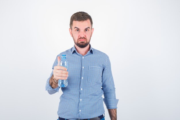 Jeune homme tenant une bouteille d'eau en plastique en chemise, jeans et à la recherche de sérieux. vue de face.