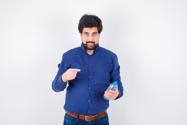 Jeune homme tenant une bouteille d'eau et la pointant en chemise bleue et en jean et l'air optimiste. vue de face.