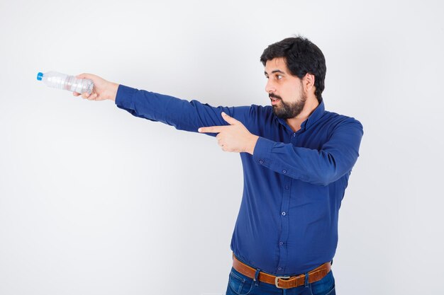 Jeune homme tenant une bouteille d'eau et pointant vers la gauche en chemise bleue et jeans et à l'optimisme. vue de face.