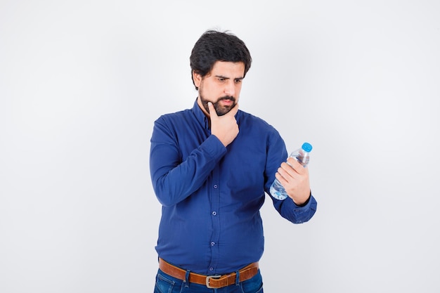 Jeune homme tenant une bouteille d'eau et tenant la main sur le menton en chemise bleue et jeans et regardant concentré, vue de face.