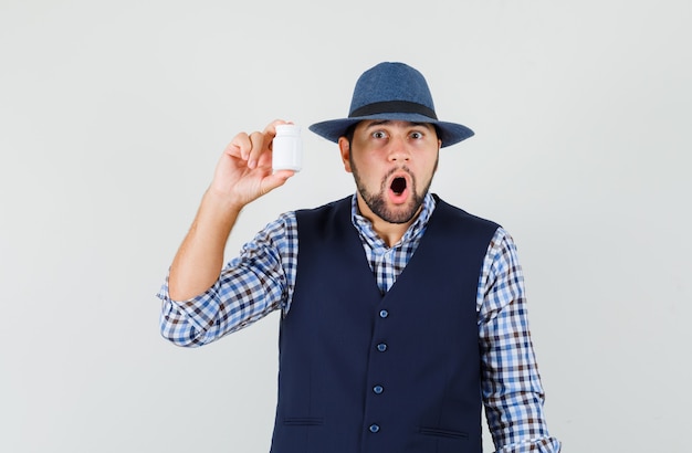 Jeune homme tenant une bouteille de pilules en chemise, gilet, chapeau et à la choqué. vue de face.