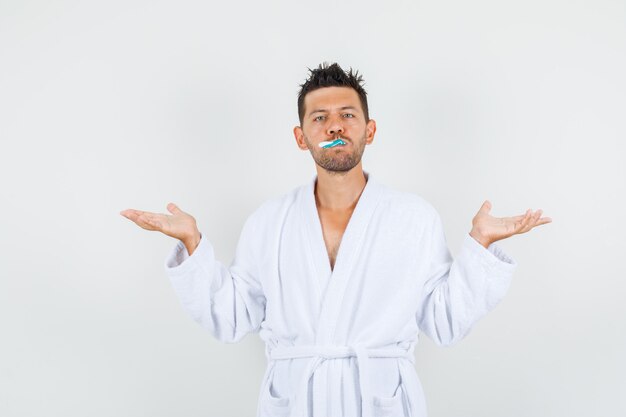 Photo gratuite jeune homme tenant une brosse à dents dans la bouche avec un geste impuissant en peignoir blanc, vue de face.