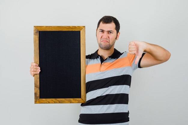 Jeune homme tenant un cadre noir tout en montrant le pouce vers le bas en t-shirt et à la mécontentement.