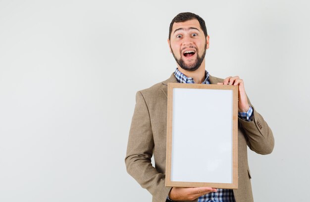 Jeune homme tenant un cadre vierge en chemise, veste et air heureux. vue de face.