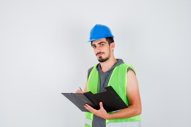 Jeune homme tenant un cahier et écrivant quelque chose dessus avec un stylo, regardant la caméra en uniforme de construction et ayant l'air heureux