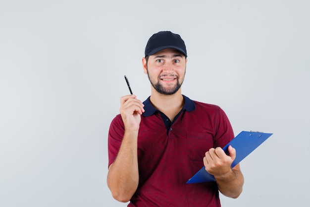 Jeune homme tenant un cahier et un stylo en t-shirt rouge, casquette noire et à la recherche intelligente, vue de face.