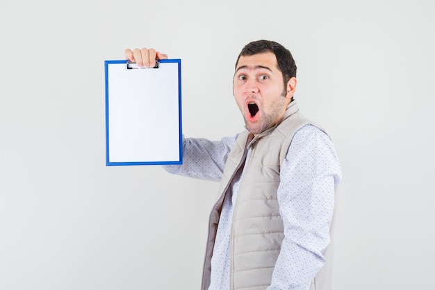 Jeune homme tenant un cahier tout en ouvrant la bouche en veste beige et à la surprise, vue de face.