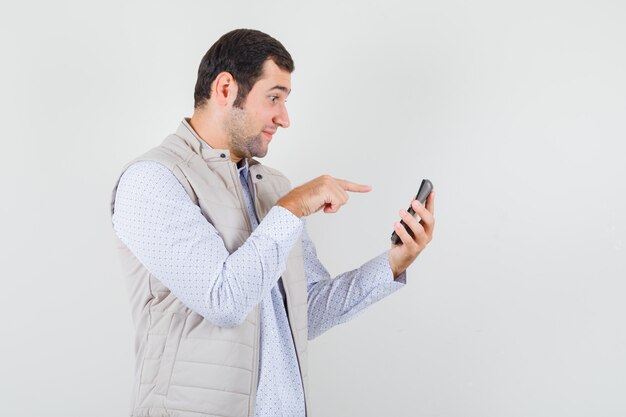 Jeune homme tenant la calculatrice dans une main et pointant vers elle avec l'index en veste beige et casquette et à la recherche de sérieux. vue de face.