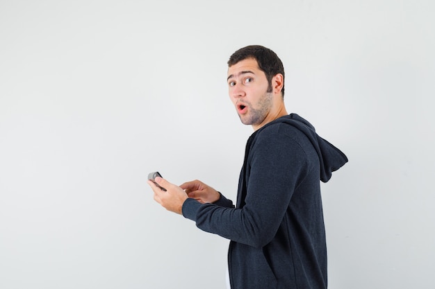 Jeune homme tenant la calculatrice et faisant quelques opérations dessus en t-shirt blanc et sweat à capuche noir à glissière sur le devant et à la surprise, vue de face.