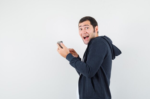 Jeune homme tenant la calculatrice et faisant quelques opérations dessus en t-shirt blanc et sweat à capuche noir à glissière sur le devant et à la surprise, vue de face.